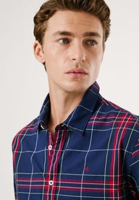 Camisa de cuadros azul marino con patrones de rejilla en rojo y blanco, cuello abotonado y pequeño logotipo bordado en el pecho. Material de algodón.
