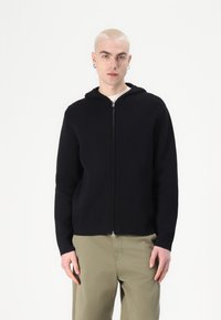 Svart zip-up hoodie tillverkad av ett mjukt tyg, med huva och långa ärmar, bärs över en ljusfärgad skjorta och matchas med kaki-byxor.