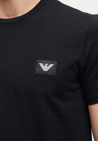 Μαύρο πουκάμισο με κοντά μανίκια και ένα μικρό τετράγωνο λογότυπο με λευκό στυλιζαρισμένο αετό στο αριστερό μέρος του στήθους.