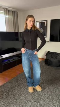 Juoda marškinėliai su sagomis, šviesiai mėlynos spalvos džinsai su laisvu kirpimu ir smėlio spalvos bateliai be raištelių. Stovi ant tekstūruoto pilko kilimo patalpose.