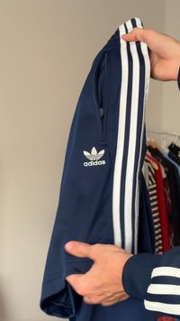 Marineblå jakke med hvit adidas-logo, med glidelåslomme og tre hvite striper langs ermene. Glatt, lett stoff.