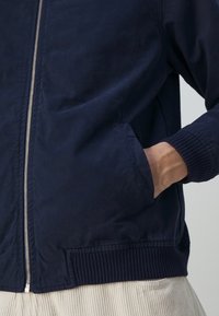 Main dans la poche d'une veste zippée bleu marine avec poignets et ourlet côtelés, portée sur un pantalon beige clair en velours côtelé.