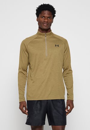 Homme portant un haut à manches longues olive avec fermeture éclair au quart et logo Under Armour ainsi qu'un short de sport texturé noir, debout devant un fond uni.