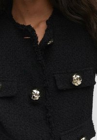 Chaqueta negra texturizada con bordes deshilachados y grandes botones plateados brillantes en el frente y los bolsillos, llevada por una persona con el cabello largo.