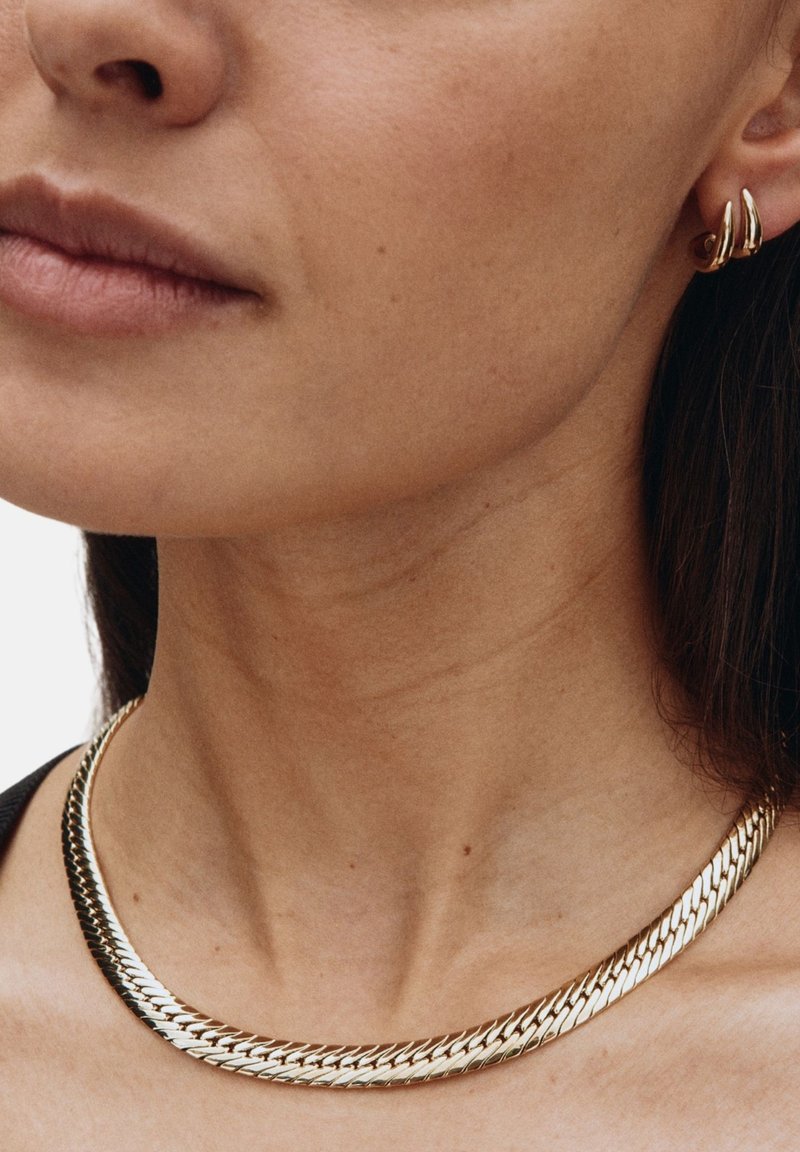 Collana in metallo color oro con un design a catena piatta e intrecciata. Presenta una superficie liscia e lucida con uno stile minimalista. Orecchini a cerchio in abbinamento.