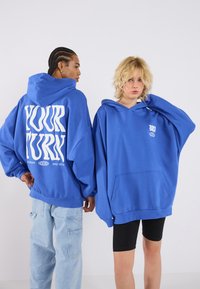 Felpe con cappuccio blu oversize con tasche frontali e grafiche: "IL TUO TURNO" in bianco bold sul retro e un piccolo logo sul davanti.