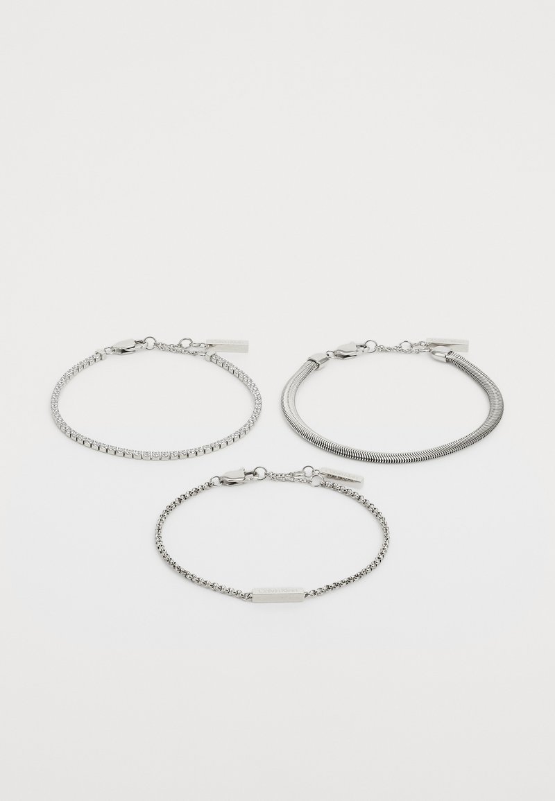 Três pulseiras de prata: uma com elos e strass, uma com um design liso e flexível, e uma com uma corrente texturizada.
