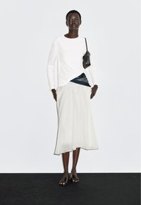 Hvid bluse med lange ærmer kombineret med en let beige nederdel. Et blåt bandana-accentsyn dukker frem, suppleret med en lille sort håndtaske og brune sandaler.