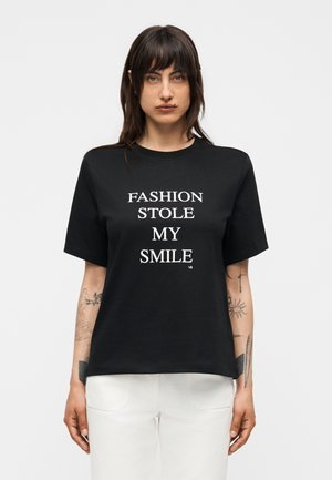 Kvinde med mørkt hår og tatoveringer iført sort t-shirt med teksten "FASHION STOLE MY SMILE" kombineret med hvide bukser mod ensfarvet baggrund.