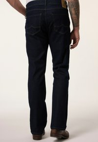 Tummansiniset farkut suoran istuvuuden kanssa, joissa on kaksi takataskua ja vaaleanruskea nahkapaikka vyötäröllä, yhdistettynä ruskeisiin saappaisiin.