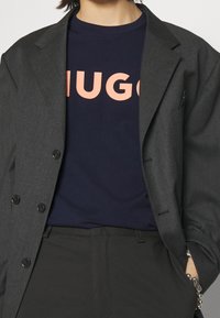 Harmaa bleiseri tummansinisen T-paidan päällä, jossa on voimakas koralli "HUGO" -logo, mustat housut ja hopeaketju. Suorat pinnat ja muotonsa mukaiset leikkaukset.