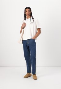 White cotton t-shirt with a graphic print, blue denim jeans, and tan sneakers; model holding a beige tote bag and standing against a white background.