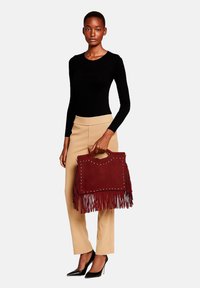Bordeaux franje handtas met studaccenten, gemaakt van suede, rechthoekige vorm, gedragen door een model met een zwart overhemd met lange mouwen en beige broek.