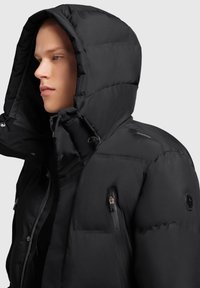 Svart puffjacka med huva, med en slät yta, dragkedjefickor och en cirkulär logodetalj på axeln.