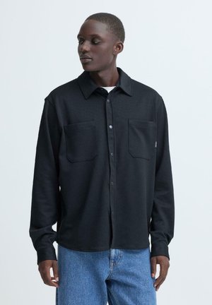 Sort skjorte med knapper, tekstureret finish, to brystlommer og klassisk krave, kombineret med lyseblå jeans.