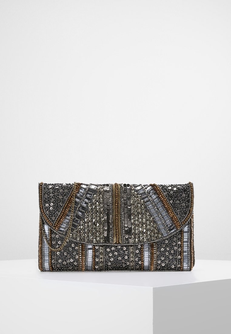 Pochette rectangulaire ornée de motifs complexes en perles et sequins argentés, noirs et dorés, avec une fine chaîne dorée, posée sur un socle blanc.
