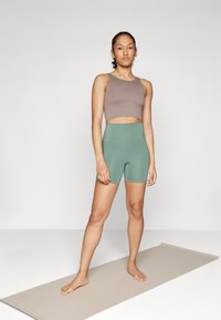 Haut court côtelé marron et short vert taille haute, tous deux en tissu extensible. Debout sur un tapis de yoga beige clair, pieds nus, arrière-plan neutre.