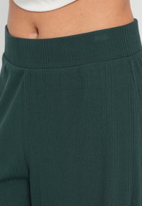 Donkergroene ribbroek met een brede, elastische tailleband en een gestructureerde stof. Voorzien van subtiele plooitjes langs de zijkanten voor extra detail.