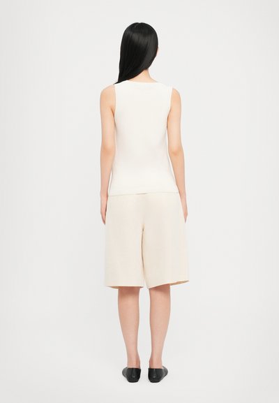 Lys beige ermeløs topp med en jevn tekstur kombinert med kremfargede vide shorts. Sorte slip-on sko. Enkel, moderne design.