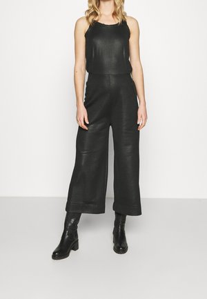 Femme portant un haut noir sans manches, un pantalon taille haute à jambes larges et des bottines noires à talons sur fond blanc uni.
