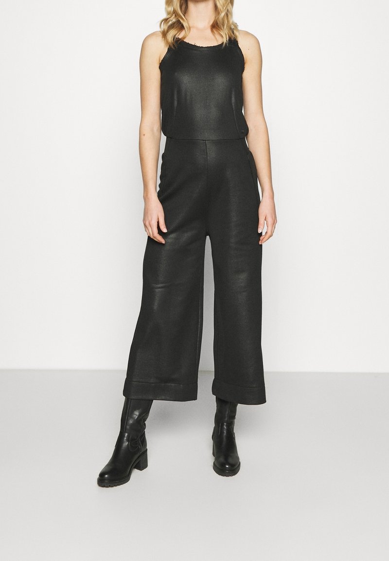 Femme portant un haut noir sans manches, un pantalon taille haute à jambes larges et des bottines noires à talons sur fond blanc uni.