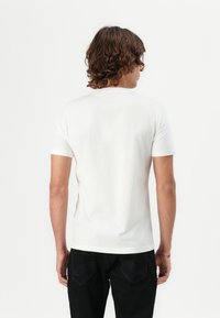 Camiseta de algodón blanca de manga corta, con cuello redondo y una textura suave. Se lleva con pantalones negros que presentan detalles de costura sutiles.