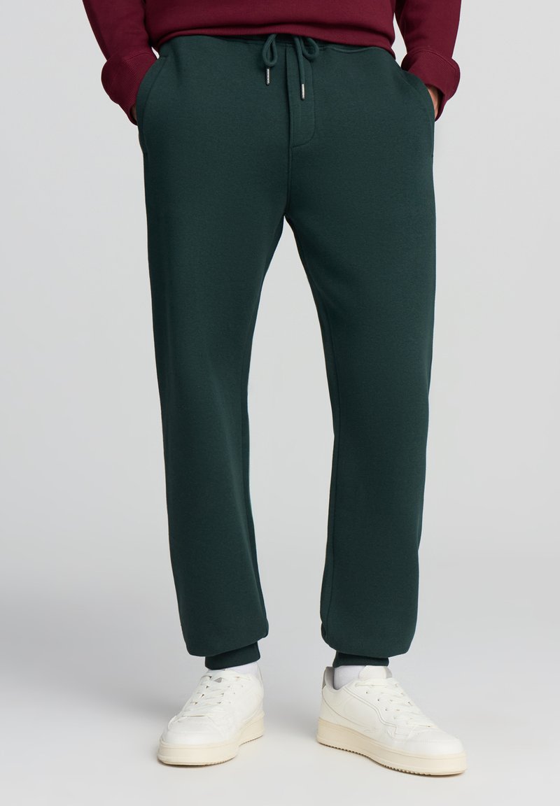 Ciemnozielone dresy z elastycznym pasem, bocznymi kieszeniami i ściągaczami. Wykonane z miękkiego materiału, charakteryzują się gładką teksturą.