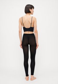 Svart bralette med tynne stropper paret med gjennomsiktige svarte leggings. Leggingsene har en glatt tekstur og en tettsittende design som fremhever silhuetten.