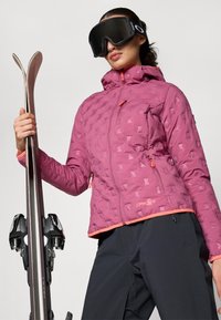 Μωβ ναυλον τζάκετ με γεωμετρικό σχέδιο, με φερμουάρ στις τσέπες και πορτοκαλί λεπτομέρειες, που συνδυάζεται με μαύρο παντελόνι σκι και κρατάει σκι.