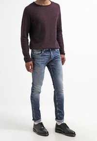 Man i en vinröd långärmad tröja, urtvättade blå slim jeans uppvikta vid ärmarna, vita strumpor och svarta dressskor.