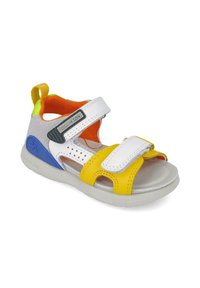 Sandału infantil colorido que presenta material sintético blanco y amarillo, correas de velcro, detalles recortados y una suela gris texturizada.