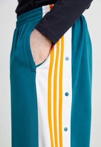 Kopparfärgade och vita träningsbyxor med oranga ränder och tryckknappar längs sidan. Har en sidoficka och elastisk midja.