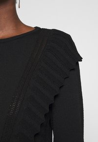 Pull noir en maille avec un motif côtelé et un accent ondulé volanté le long de la couture de l'épaule, avec un col rond.
