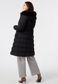 Zwarte pufferjas met een nepbont capuchon, gequilt design, elastische taille en zijzakken, gecombineerd met grijze wijde broek en laarzen met luipaardprint.