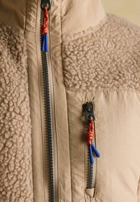 Beige fleece jacket with two black zippers, each featuring a red and yellow cord pull with blue plastic ends.