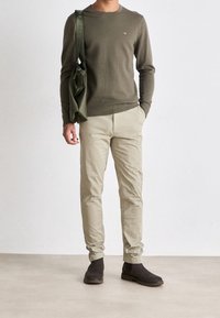 Grüner Strickpullover mit gerippten Bündchen, beigefarbene Slim-Fit-Hose und dunkelbraune Stiefeletten. Enthält eine grüne Schultertasche mit Riemen.