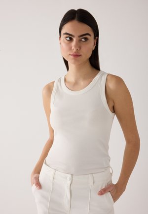 Jonge vrouw met donker haar, gekleed in een wit mouwloos geribd shirt en witte broek, handen in de zakken, kijkt opzij tegen een effen achtergrond.