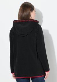 Veste à capuche noire avec broderie florale rouge le long du bord de la capuche, dotée de manches longues et de finitions rouges aux poignets et à l'ourlet.