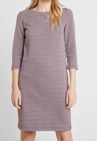 Femme portant une robe mi-longue à manches longues avec un motif pied-de-poule rouge, noir et blanc, debout devant un fond uni.