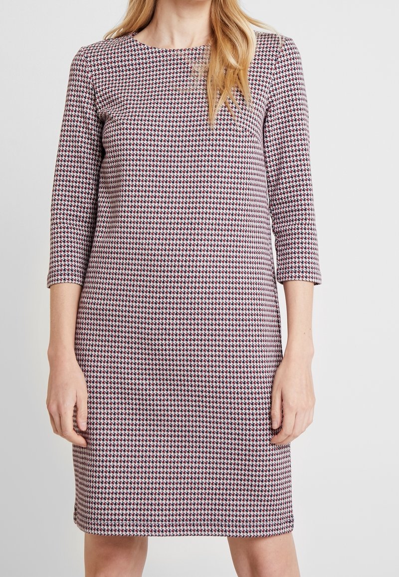 Femme portant une robe mi-longue à manches longues avec un motif pied-de-poule rouge, noir et blanc, debout devant un fond uni.