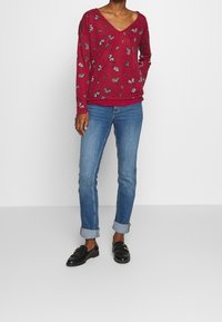 Röd långärmad V-ringad tröja med blommig fjärilsdesign, kombinerad med ljusblå uppvikta jeans och svarta loafers.