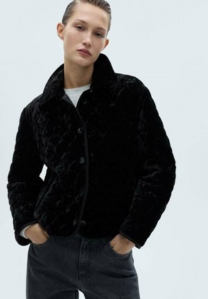 Veste matelassée noire avec une texture douce et moelleuse. Présente un col, une fermeture à boutons et des finitions contrastantes. Assortie avec un jean foncé.