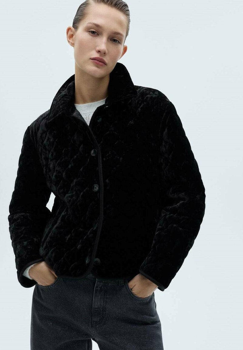 Sorte quilted jakke med en blød, luksuriøs tekstur. Har en krave, knaplukning og kontrasterende kant. Båret sammen med mørke jeans.