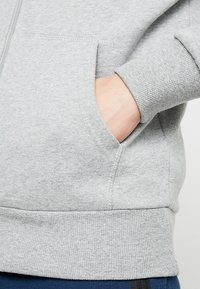 Ruka spočívající v přední kapse světle šedé mikiny s žebrovanými manžetami a lemem, na bílém pozadí.