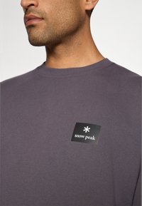 Camicia a maniche lunghe viola con colletto a girocollo a costine. Presenta un'etichetta nera con testo e logo bianchi. Tessuto morbido e leggero.