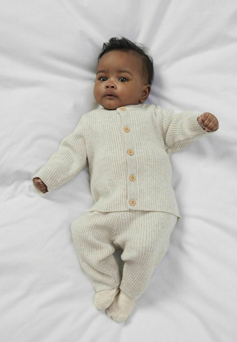 Elfenbensfärgad ribbad stickad babyoutfit med en knäppt topp med lång ärm och matchande byxor, visad mot en vit bakgrund.