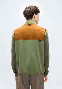 Ανδρικό μπου φάν με πράσινο σώμα και καφέ επάνω μέρος, με ομαλή υφή, ψηλό γιακά και μικρή διακριτική λογότυπο.