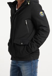Veste softshell noire avec une poche poitrine zippée, des poignets élastiques et un ourlet resserré ; dotée d'un écusson logo sur la manche gauche.