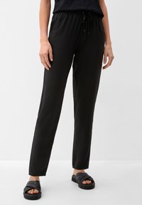 Pantalons noirs, légers, avec taille élastique et lien de serrage. Texture lisse et coupe fuselée, associés à des sandales à glissière noires.