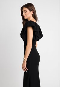 Czarna sukienka o dopasowanej sylwetce, z dużymi falbaniastymi rękawami i miękką tkaniną. Uzupełniona złotym zegarkiem na nagim nadgarstku.